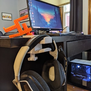 May include: A 3D printed orange headset stand with a black and white headset hanging on it. The stand is attached to a black desk with a computer monitor in the background.