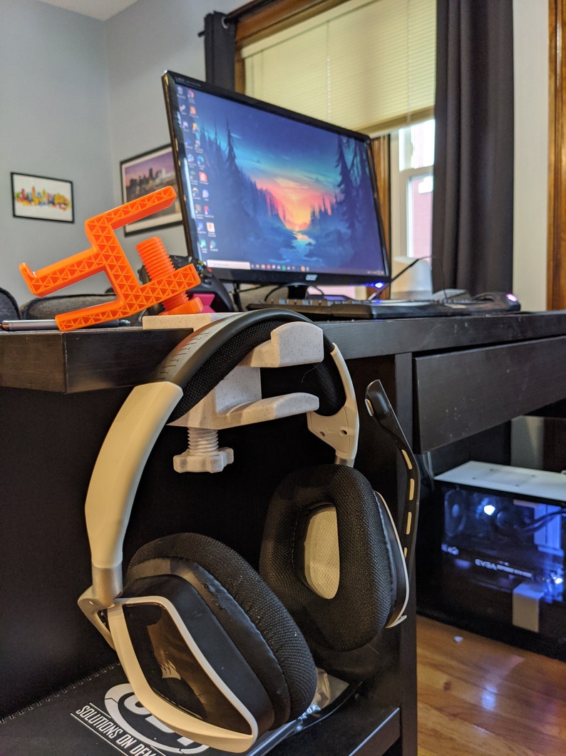 May include: A 3D printed orange headset stand with a black and white headset hanging on it. The stand is attached to a black desk with a computer monitor in the background.