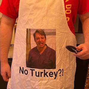 Peut inclure: Tablier blanc avec une image d'un homme avec une expression surprise et le texte "No Turkey?!" imprimé dessus. Le tablier a du texte jaune sur un t-shirt rouge en dessous.