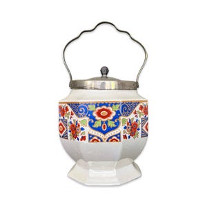 May include: A white ceramic jar with a silver lid and handle. The jar features a colorful floral pattern in red, blue, and gold. The jar has a pedestal base and a silver handle for carrying.