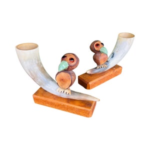 May include: Two wooden bird sculptures with large eyes and green beaks are perched on top of two wooden bases. Each base is attached to a curved, light brown horn.