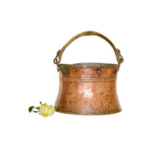 May include: A copper bucket with a brass handle. The bucket has a hammered texture and a worn patina. A single white rose is in the foreground.