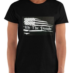 Puede incluir: Camiseta negra con una bandera estadounidense blanca desgastada y el texto "We The People" impreso en ella.