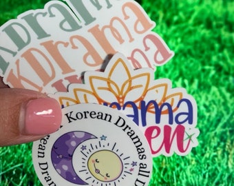 May include: Three stickers with a Korean drama theme. The first sticker is pink and white with the word "drama" written in a cursive font. The second sticker is a yellow and orange sun with a flower design. The third sticker is a circle with a purple moon and a yellow sun with the text "Korean Dramas All Day All Night".