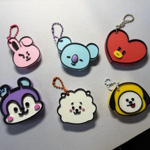 Op de afbeelding: Zes kleurrijke sleutelhangers met karakters. De sleutelhangers tonen een roze konijn, een lichtblauwe koala, een rood hart, een paars eekhoorntje, een wit schaap en een gele hond. Elke sleutelhanger heeft een metalen ketting.