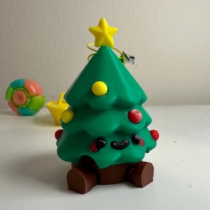 May include: A green Christmas tree ornament with a yellow star. The tree has red and yellow baubles and a brown base. A colourful ball and yellow boat are in the background.