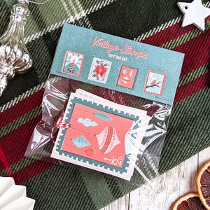 Pu&ograve; includere: Un set di etichette regalo di Natale vintage con sfondo blu e bianco e illustrazioni rosse e bianche. Le etichette raffigurano una candela, una stella di Natale, un ornamento natalizio e un uccello. Le etichette sono confezionate in un sacchetto di plastica trasparente e includono il testo "Vintage Stamps Gift Tag Set".