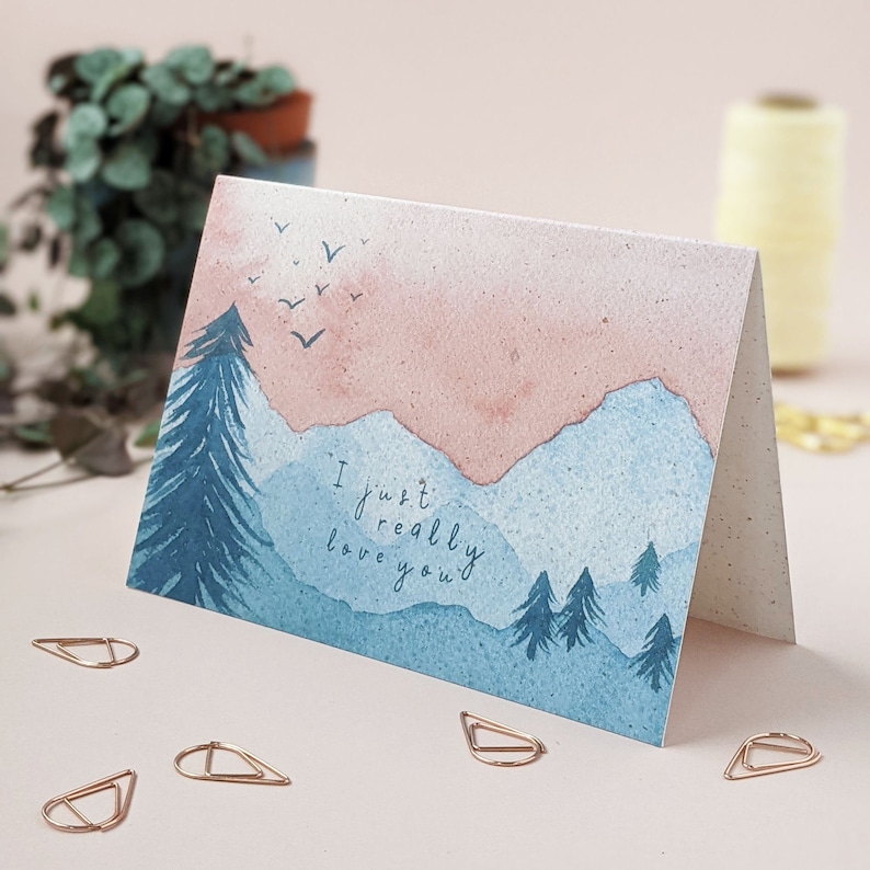 Op de afbeelding: Een aquarel-groetkaart met een blauw en roze berglandschap en de tekst "Ik hou echt van je".