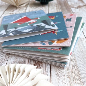 May include: A stack of colorful Christmas cards with illustrations of Santa Claus, a snowman, and other winter scenes. The cards are made of recycled paper and have a rustic, handmade feel.