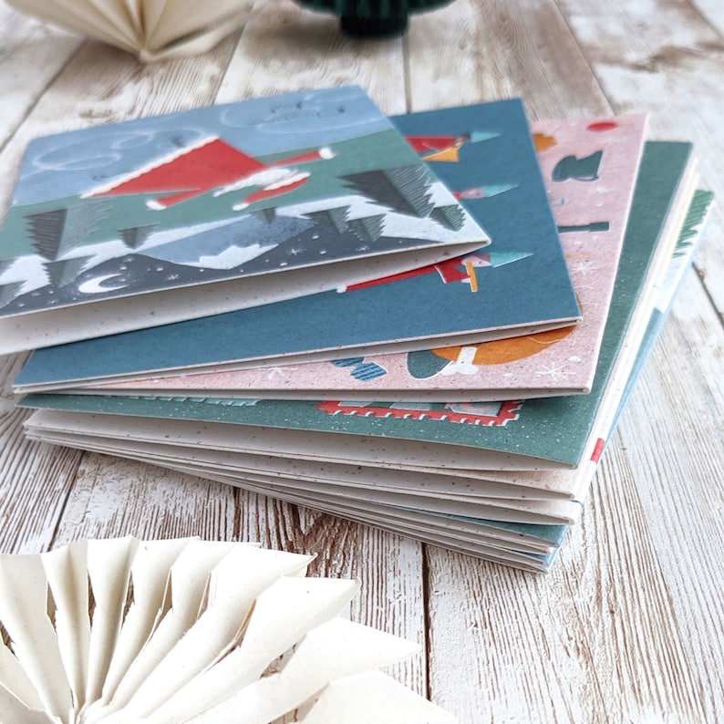 May include: A stack of colorful Christmas cards with illustrations of Santa Claus, a snowman, and other winter scenes. The cards are made of recycled paper and have a rustic, handmade feel.