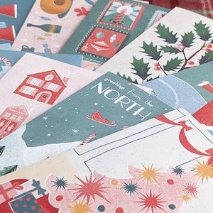 May include: A stack of colorful Christmas cards with various illustrations, including a house, a Christmas tree, and a holly sprig. The top card features the text "Greetings from the North".