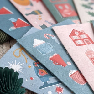 May include: A stack of blue and pink cards with illustrations of winter scenes. The cards feature a cup of hot chocolate with whipped cream, a house with a chimney, and a pair of ice skates.