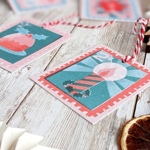 Pu&ograve; includere: Due etichette regalo di Natale con un bordo rosa e rosso. Un'etichetta presenta una candela a strisce rosso e bianco con una fiamma e agrifoglio. L'altra etichetta presenta un budino di Natale rosso e bianco con agrifoglio.