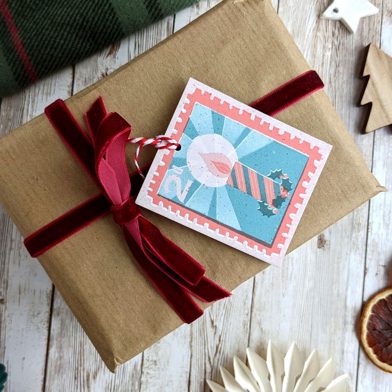 Pu&ograve; includere: Un pacchetto regalo in carta marrone legato con un nastro di velluto rosso e un'etichetta regalo a tema natalizio con il numero "2" e un'illustrazione di una candela.