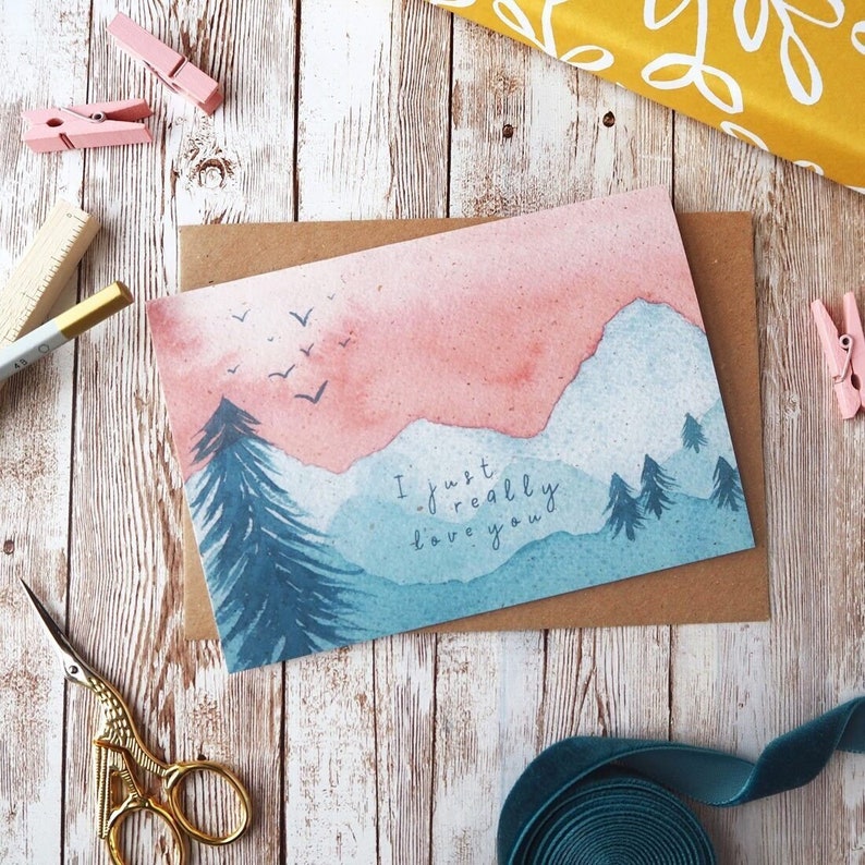 Op de afbeelding: Een aquarelkaart met een berglandschap en de tekst "I just really love you". De bergen zijn geschilderd in tinten blauw en roze, met een paar vogels die erboven vliegen. De kaart is perfect voor een romantische gelegenheid.