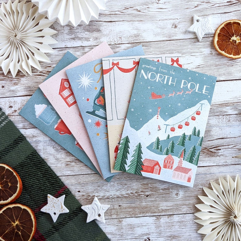 May include: Set of five Christmas cards featuring a winter scene with a ski lift, snow-covered mountains, and houses. The cards are printed on recycled paper and have a rustic, vintage feel. The text on the front of the card reads "Greetings from the North Pole".