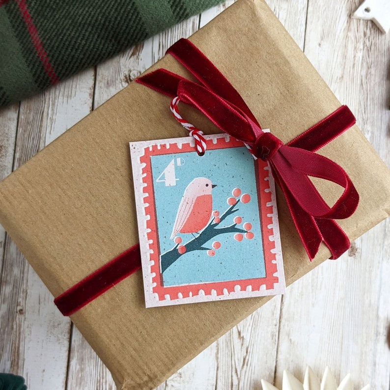 Pu&ograve; includere: Un sacchetto regalo di carta marrone con un nastro di velluto rosso legato intorno. Un'etichetta regalo rossa e bianca con un pettirosso su un ramo e il testo "4d" &egrave; attaccato al nastro.