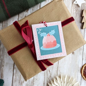 Pu&ograve; includere: Una scatola regalo di carta marrone con un nastro di velluto rosso legato intorno. Un'etichetta regalo di budino di Natale rosa e verde &egrave; attaccata al nastro. L'etichetta dice "2d" nell'angolo.