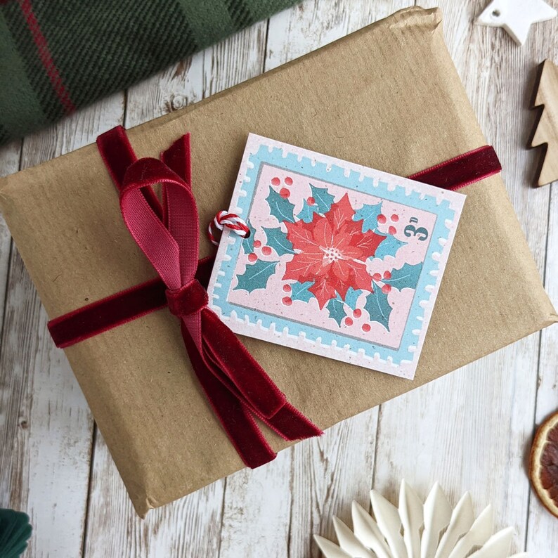 Pu&ograve; includere: Un sacchetto regalo di carta marrone con un nastro di velluto rosso e un'etichetta regalo a tema natalizio. L'etichetta presenta un fiore di stella di Natale con foglie verdi e bacche rosse su uno sfondo rosa con un bordo blu. L'etichetta ha anche il numero "3" stampato su di essa.