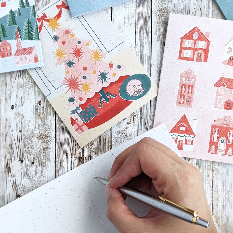 May include: A hand writing on a blank white card with a pen. The card is on a wooden surface with a colorful Christmas card featuring a Christmas tree and houses in the background.