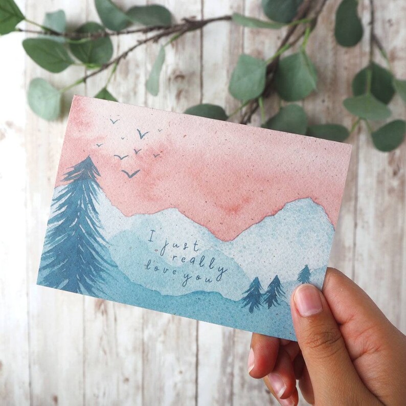 Op de afbeelding: Een aquarelkaart met een roze en blauwe berglandschap. De kaart heeft de tekst "I just really love you" erop.