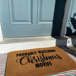以下が含まれることがあります： 黒い筆記体で「Probably Watching Christmas Movies」と書かれた茶色のココナッツ製のドアマットです。