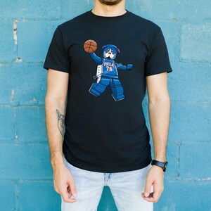 Peut inclure: T-shirt noir avec un ours bleu de dessin animé tenant un ballon de basket. L'ours est conçu dans un style Lego et porte un maillot avec "PHILA 76" et le numéro 76. L'ours a un visage blanc et des oreilles bleues.