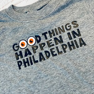 Puede incluir: Una camiseta gris de cuello redondo con la frase "GOOD THINGS HAPPEN IN PHILADELPHIA" impresa en negro y naranja. La camiseta presenta un gráfico de dos ojos naranjas y blancos encima de las palabras.