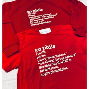 Może przedstawiać: Czerwony T-shirt z białym tekstem, który brzmi "go phils /'go fils/ phrase used to mean "believe," "rise above," "let's go fighting!" See also: ring that bell or hit that jawn. origin: philadelphia"