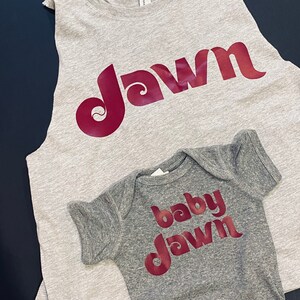 Peut inclure: Un d&eacute;bardeur gris sans manches et un body b&eacute;b&eacute; gris avec le mot "dawn" dans une police r&eacute;tro rouge. Le d&eacute;bardeur dit "dawn" et le body dit "baby dawn".