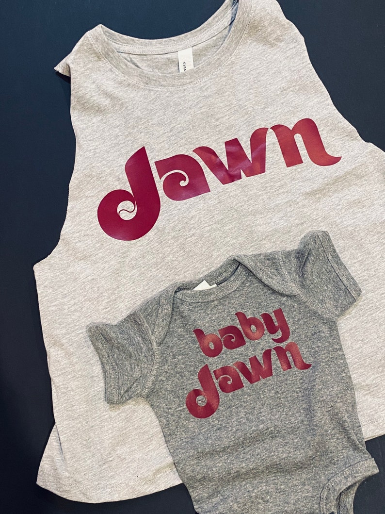 Peut inclure: Un d&eacute;bardeur gris sans manches et un body b&eacute;b&eacute; gris avec le mot "dawn" dans une police r&eacute;tro rouge. Le d&eacute;bardeur dit "dawn" et le body dit "baby dawn".