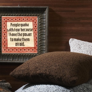 Op de afbeelding: Een ingelijst borduurwerk met de tekst "People quake with fear because I have the power to make them afraid." Het borduurwerk zit in een donkerhouten lijst met een decoratieve rand. De lijst hangt aan een donkerhouten muur. Twee bruine en grijze kussens liggen op een bank voor de lijst.