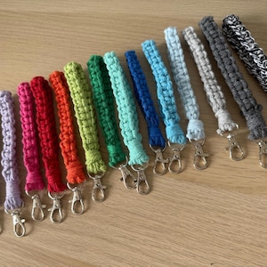 May include: A selection of macramé wrist lanyards in a spectrum of colours: purple, pink, red, orange, green, teal, blue, light blue, grey, dark grey, and black and white. Each lanyard has a silver clasp.