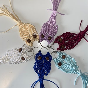 May include: A selection of handmade macrame owl keyrings in cream, white, lavender, burgundy, blue, and light blue. Each owl has wooden bead eyes and a silver keyring.