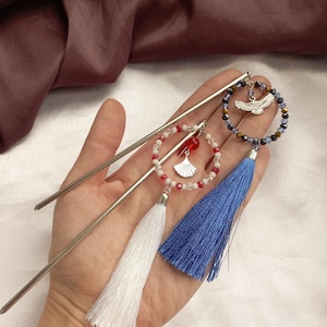 May include: A hand holds three decorative hair sticks. Two sticks are silver-toned metal. One hair stick has a white tassel and a red and white beaded circle with a fan charm. The other has a blue tassel and a beaded circle with a silver eagle charm.