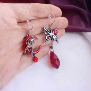 May include: Asymmetrical earrings. One earring has a red crescent moon with a silver figure and a red teardrop gem. The other has a silver butterfly with a red gem and a red teardrop gem. The earrings are held in a hand.