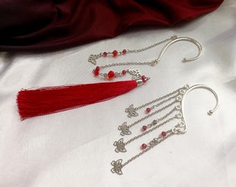 May include: Two silver-toned ear cuffs featuring delicate chains, red beads, and butterfly charms. One cuff includes a long, vibrant red tassel attached to the chain structure, while the other features multiple dangling chains with butterfly accents.