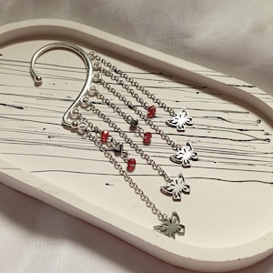 May include: Silver ear cuff with dangling chains and red beads, ending in butterfly charms. The ear cuff is on a white tray with black lines.