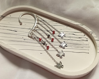 May include: Silver ear cuff with dangling chains and red beads, ending in butterfly charms. The ear cuff is on a white tray with black lines.