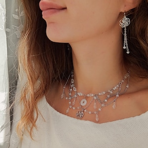 Könnte beinhalten: Silberfarbenes Halsketten- und Ohrring-Set. Die Halskette hat mehrere Stränge mit klaren Perlen und einen Rosenanhänger. Die Ohrringe haben ein Rosendesign mit hängenden Ketten und Perlen. Der Schmuck wird von einer Person getragen, die ein weißes Oberteil trägt.