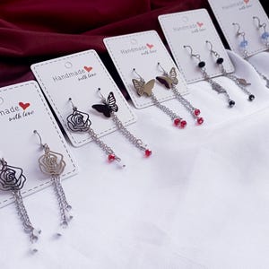 May include: A collection of handmade earrings displayed on white cards. The earrings feature various designs, including roses, butterflies, and abstract shapes, with dangling chains and decorative beads. The cards read "Handmade with love."