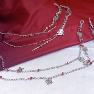 May include: Two silver-toned metal eyeglass chains featuring delicate link designs. One chain includes small white beads, a rose charm, and a sword charm. The second chain features small red beads and multiple butterfly-shaped metal charms.