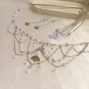May include: A silver-coloured jewellery set comprising a headband, earrings, and a hair accessory. The set features chains, blue and clear beads, and fan-shaped charms. The jewellery is displayed on a white surface.