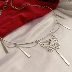 Pode incluir: Corrente de corpo prateada com um pendente de borboleta. A delicada corrente apresenta um design curvo com detalhes retangulares pendurados e um adorno central em forma de borboleta. A joia é exibida sobre um tecido vermelho e um fundo branco.
