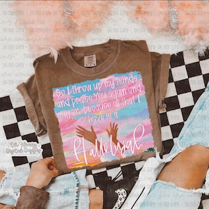 Op de afbeelding: Een bruin T-shirt met een aquarelontwerp met een roze, blauwe en witte lucht met twee opgeheven handen. De tekst "So I throw up my hands and praise You again and again because all that I have is a Hallelujah" is geschreven in wit en roze.