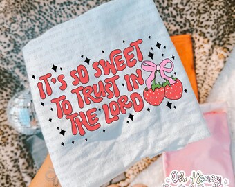 May include: A light blue t-shirt with the text "It's so sweet to trust in the Lord" in red, outlined in a darker red. The design includes two strawberries with a pink bow and black star accents. The shirt is folded.