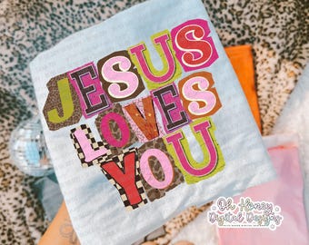 May include: Light blue t-shirt with the text "JESUS LOVES YOU" in a colorful, collage-style design. The letters are in various colors and patterns, including leopard print and checkered. The shirt is folded, with other items in the background.