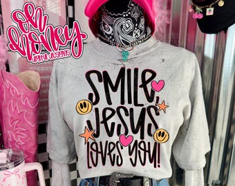 May include: A light gray sweatshirt with the text "SMILE JESUS LOVES YOU!" in black, pink, and orange. The sweatshirt is paired with blue jeans and a black belt. A pink cowboy hat and a black cap are also visible.