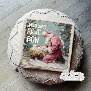 May include: A decorative pillow featuring a painting of Santa Claus kneeling before baby Jesus. The artwork is framed with gold trim and includes the text "EVERY KNEE SHALL BOW" in gold lettering. The pillow is placed on a round, neutral-toned cushion.