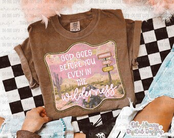 Puede incluir: Camiseta marrón con un gráfico temático del desierto. El gráfico presenta una carretera, un cactus y el texto "GOD GOES BEFORE YOU EVEN IN THE wilderness". El diseño está enmarcado en un borde rosa y dorado.